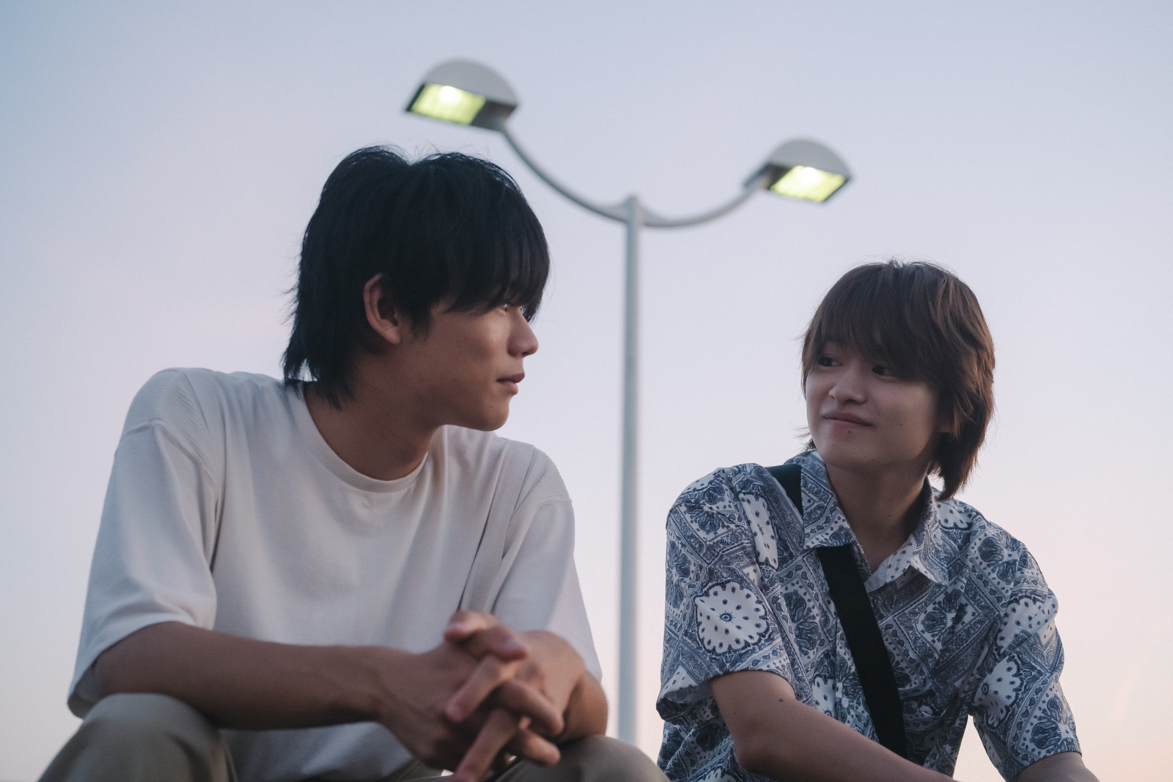 Two young men sitting side by side outdoors at dusk, smiling and looking at each other.