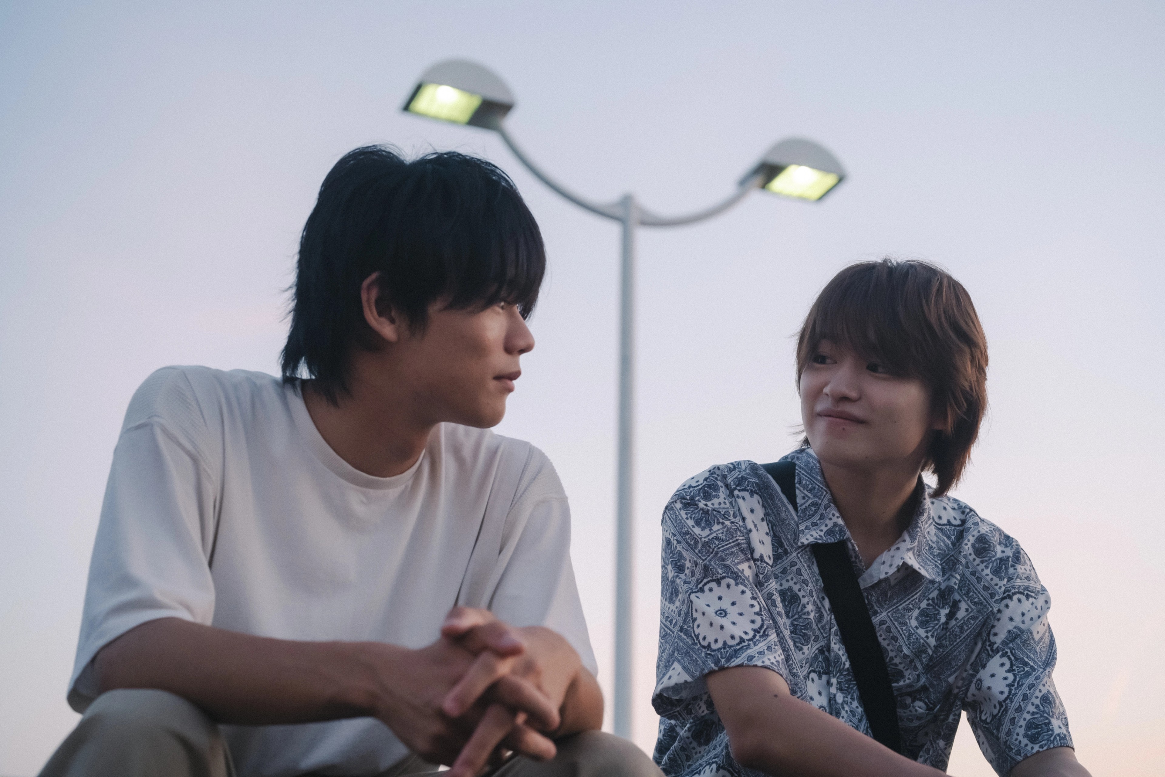 Two young men sitting side by side outdoors at dusk, smiling and looking at each other.