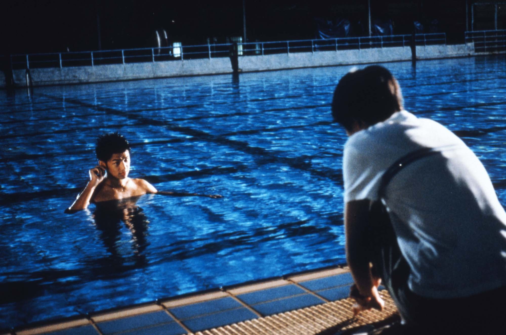 A person crouching next to a swimming pool looks at a person inside the pool.
