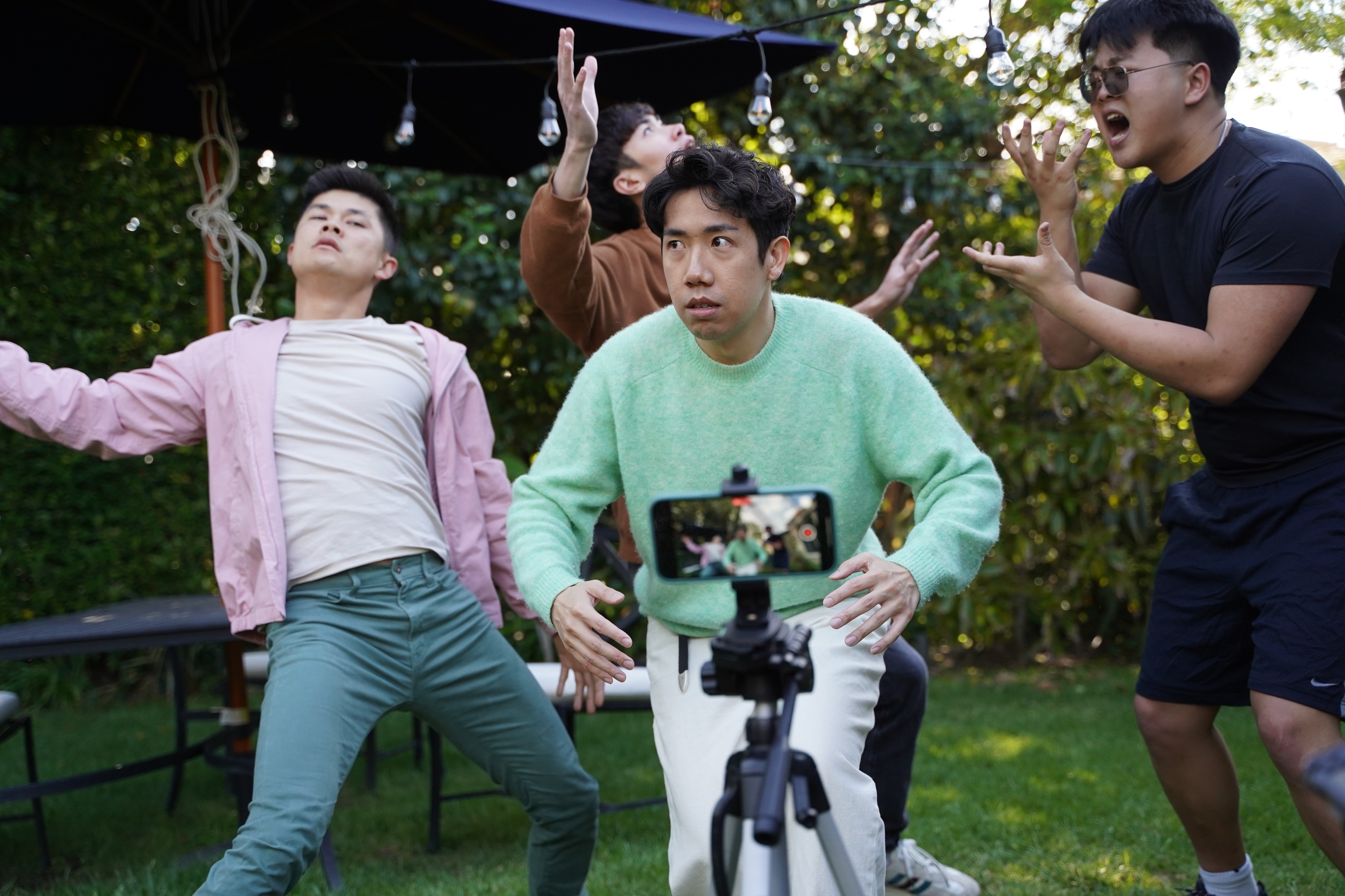 Four men performing dramatically in a garden while being filmed on a smartphone.