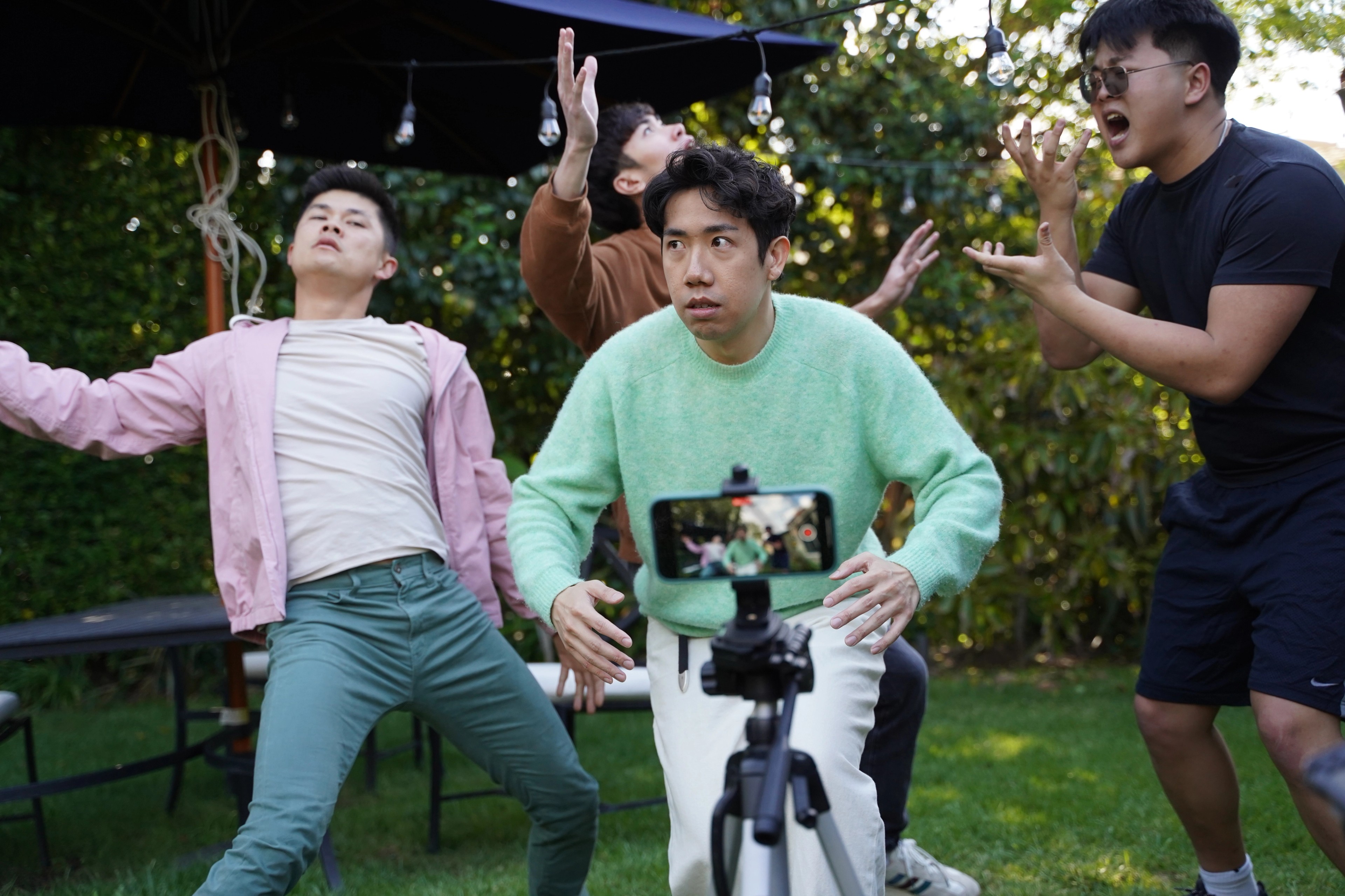 Four men performing dramatically in a garden while being filmed on a smartphone.