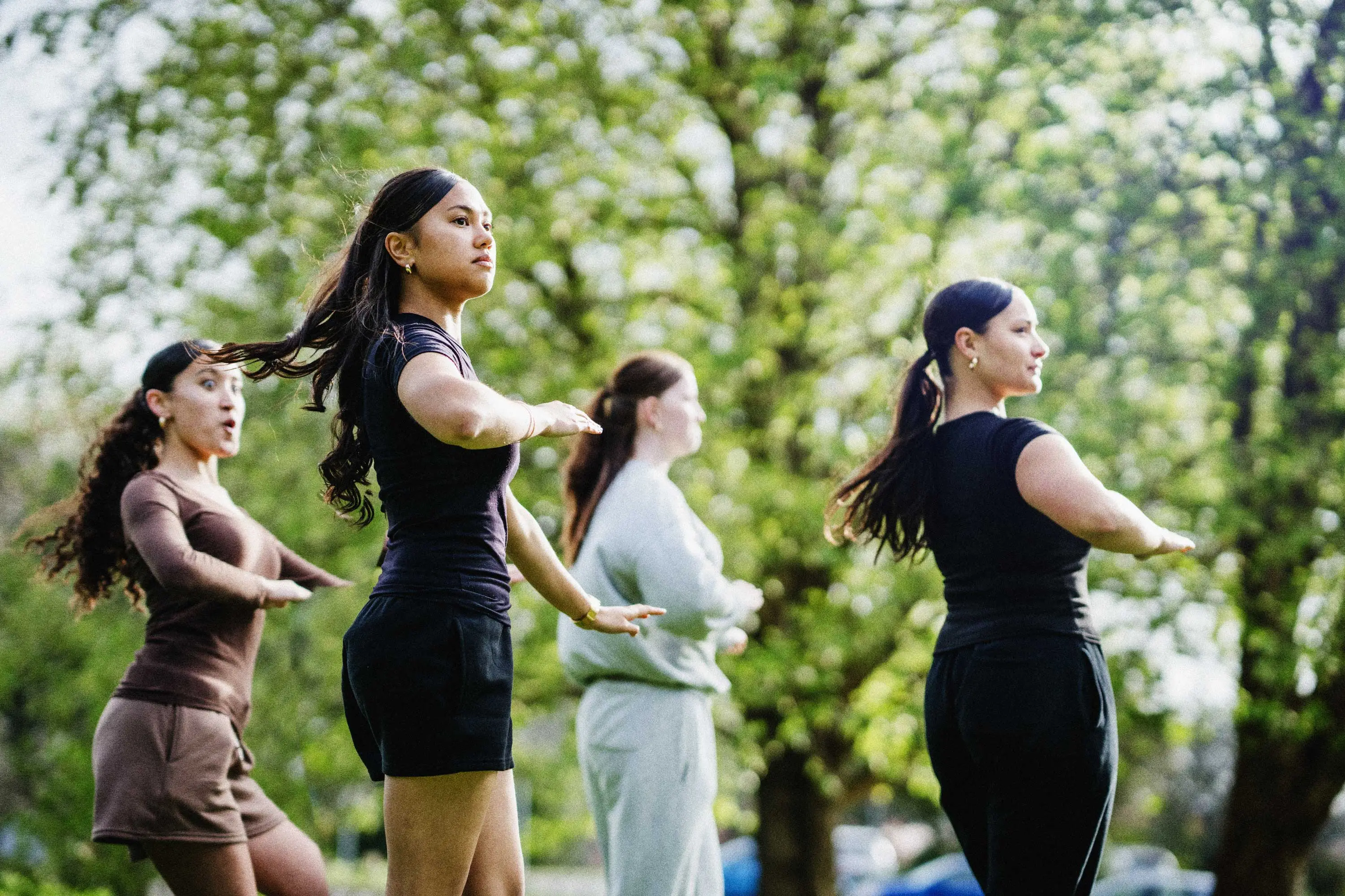 Four young women perform kapa haka outdoors against the bright green backdrop of trees.