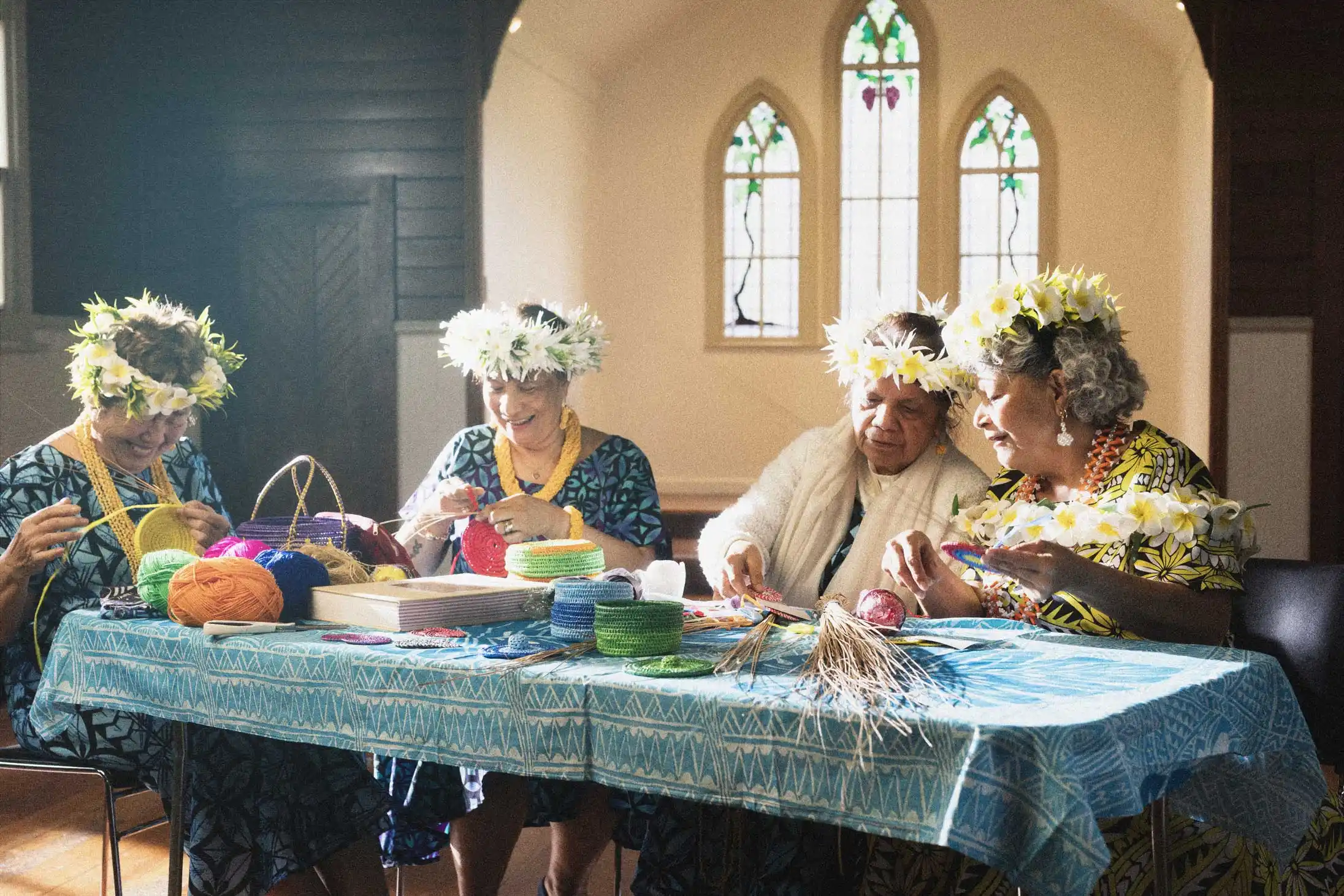 A group of older matua Niuean women sitting around a table weaving in a community hall, wearing bright dresses and fou fou / flower headdresses, surrounded by weaving craft materials.