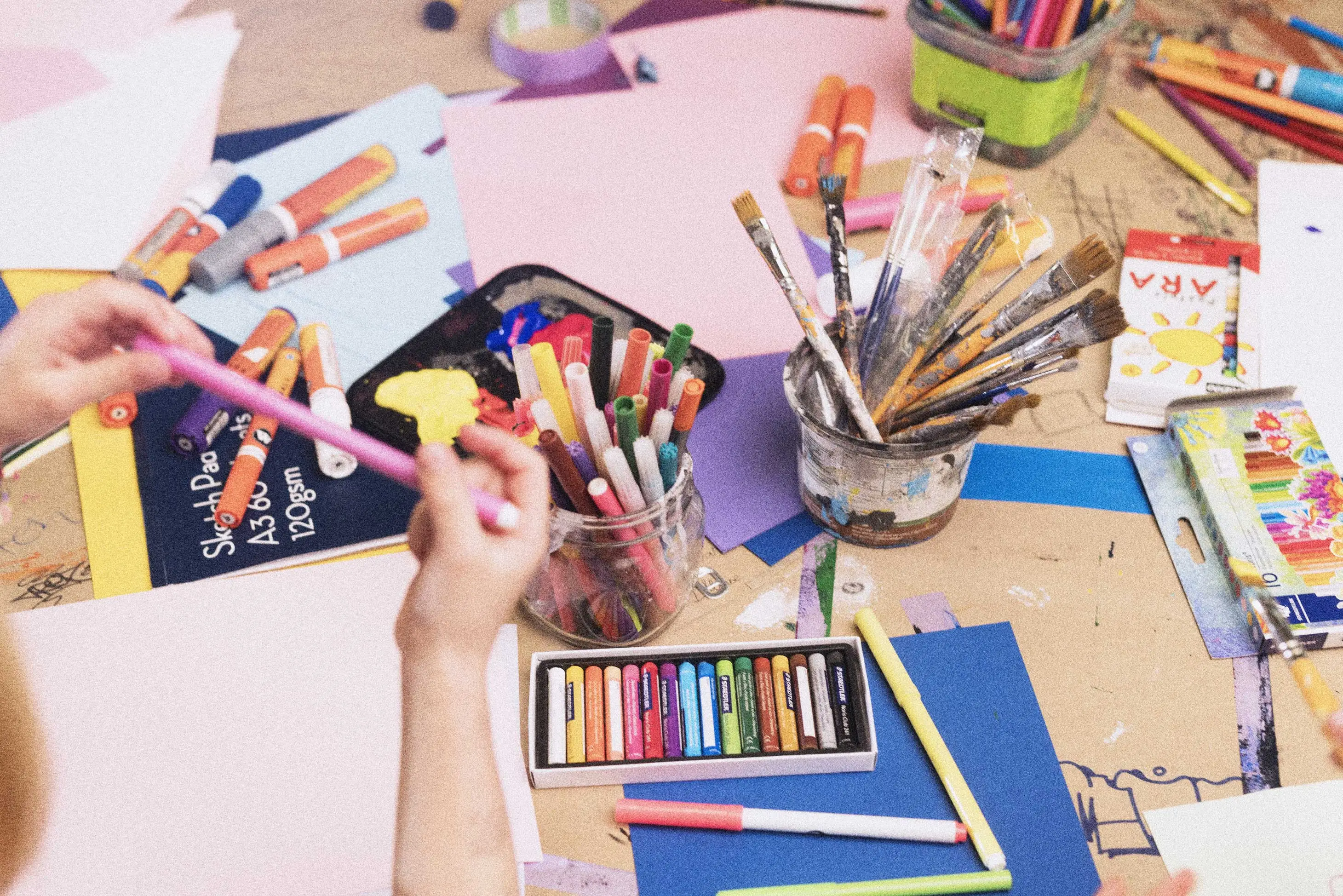 A close up image of a desk in a school art room with jars of paintbrushes, colour pens and paper scattered about.