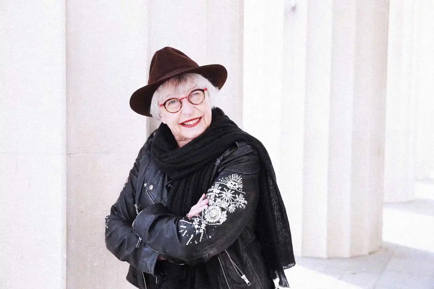 Portrait of NZ filmmaker Gaylene Preston, surrounded in bright daylight, standing in front of white columns. A dark black wool scarf around her neck, wearing a sequinned black leather jacket, glasses and a dark brown fedora hat, smiling directly to camera.