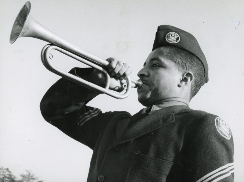 A Civilian Conservation Corps enrollee in dress uniform sounds a bugle, the CCC insignia on his cap — National Archives photo