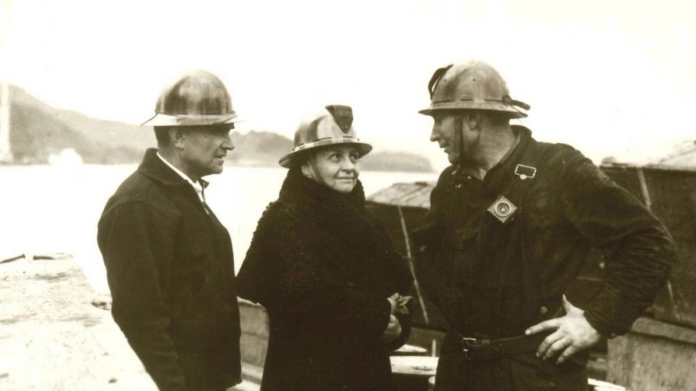 Frances Perkins, U.S. Secretary of Labor, in a hard hat speaking with two workers at a worksite