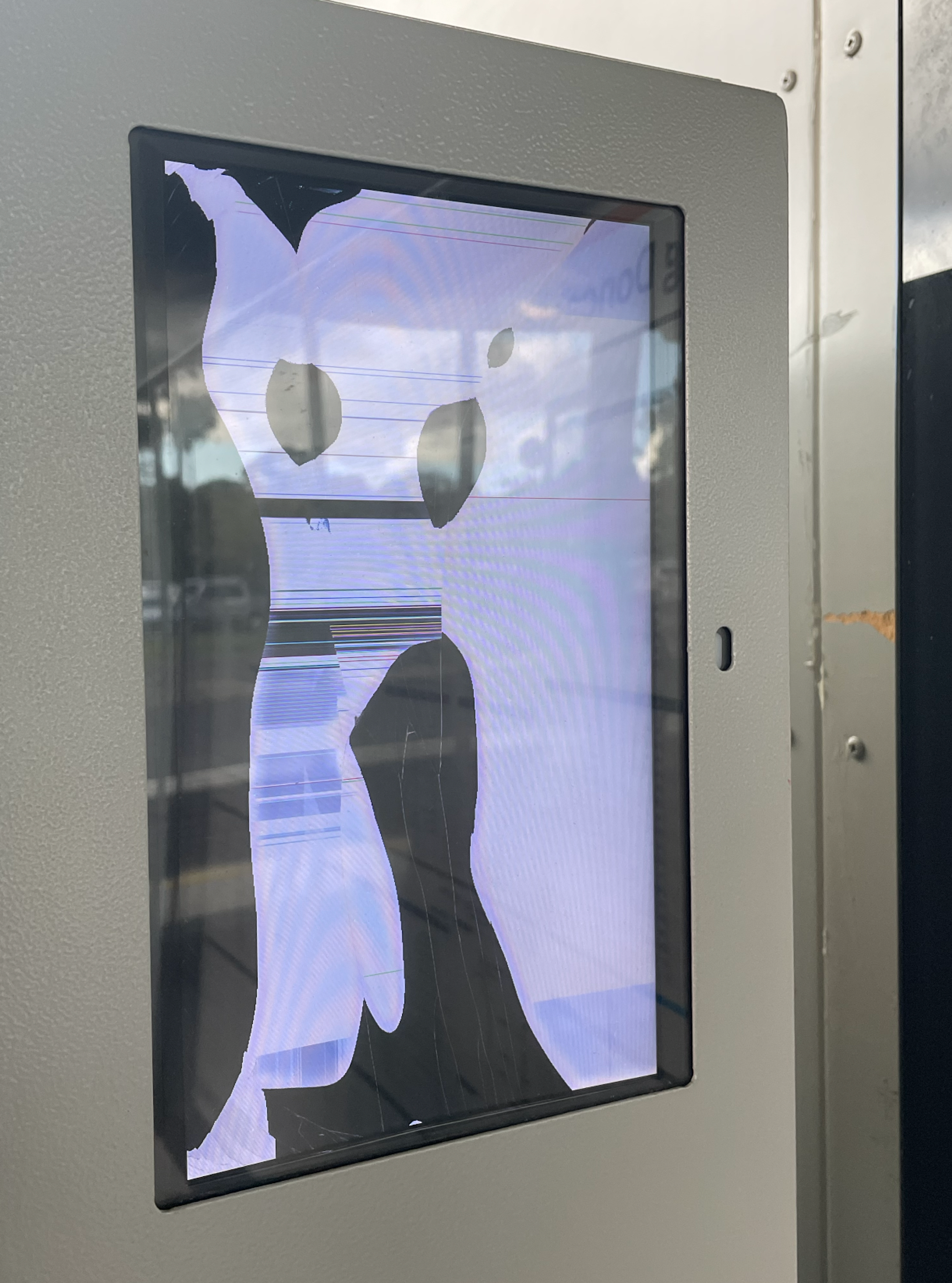 A glitched out LCD screen with a black border embedded in the top right-hand corner of a utilitarian grey vending machine. The broken screen renders a figurative character, cartoon like. The fictitious graphic character could be wearing a mask and striking a pose, ready to dance—it has hatched lines down one leg, further imperfections of the broken display. A reflection on the perspex shows a partially cloudy sky above a carpark at a bus terminal.