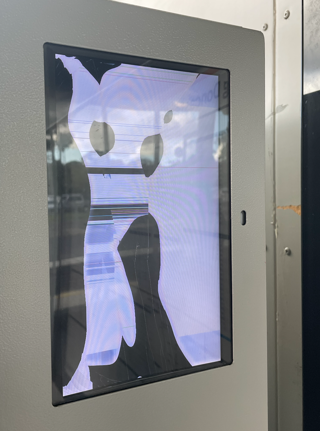 A glitched out LCD screen with a black border embedded in the top right-hand corner of a utilitarian grey vending machine. The broken screen renders a figurative character, cartoon like. The fictitious graphic character could be wearing a mask and striking a pose, ready to dance—it has hatched lines down one leg, further imperfections of the broken display. A reflection on the perspex shows a partially cloudy sky above a carpark at a bus terminal.