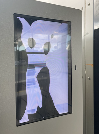 A glitched out LCD screen with a black border embedded in the top right-hand corner of a utilitarian grey vending machine. The broken screen renders a figurative character, cartoon like. The fictitious graphic character could be wearing a mask and striking a pose, ready to dance—it has hatched lines down one leg, further imperfections of the broken display. A reflection on the perspex shows a partially cloudy sky above a carpark at a bus terminal.