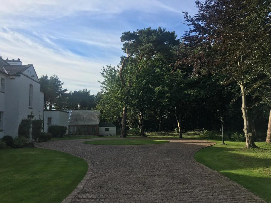 A cobbled driveway curving between lawns and mature trees towards a white house