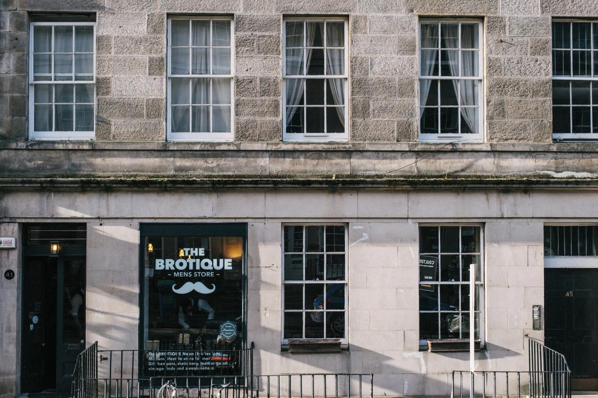 Georgian sandstone terrace with The Brotique Mens Store shopfront: a window with the shop name and a moustache graphic, black railings and a dark door