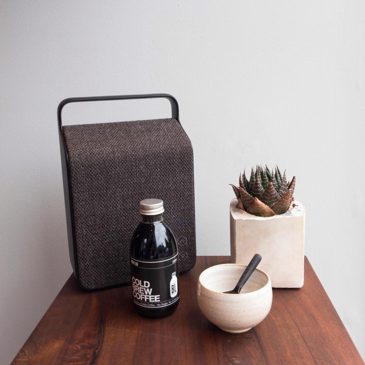 Still life on a wooden table: a fabric-covered speaker, a bottle of cold brew coffee, a succulent in a concrete pot and a white mortar and pestle