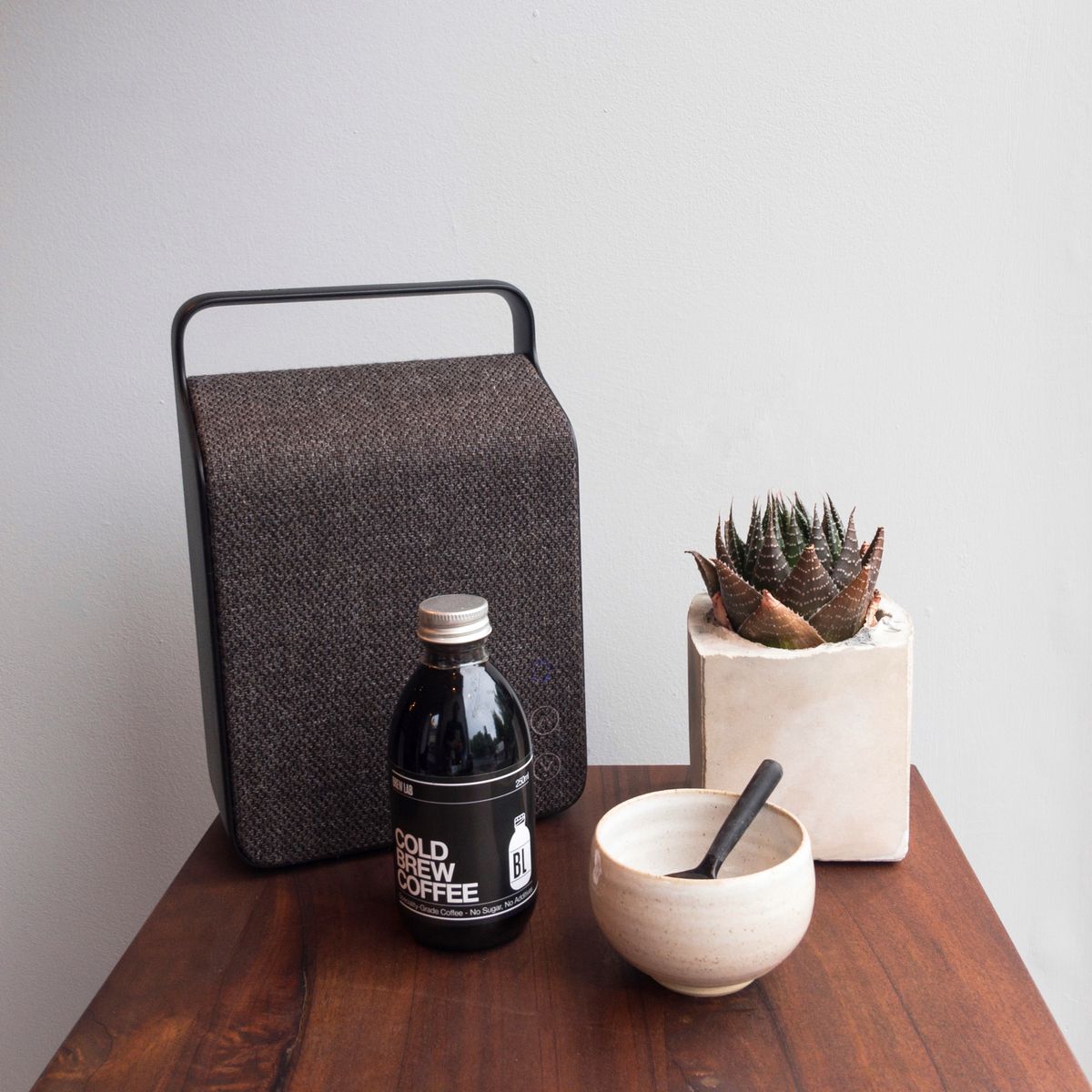 Still life on a wooden table: a fabric-covered speaker, a bottle of cold brew coffee, a succulent in a concrete pot and a white mortar and pestle