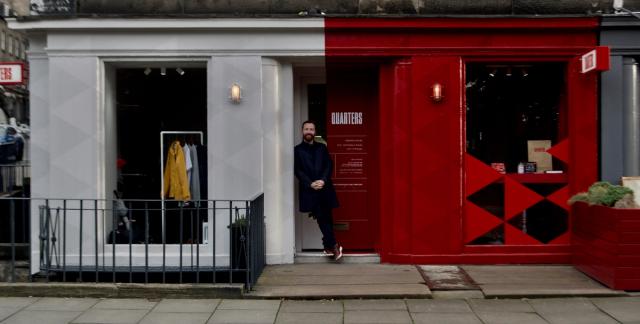 Wide view of the Quarters shopfront: a white-painted bay with a window display of clothing beside a red bay with a large red-and-black diamond graphic, and a man in the doorway