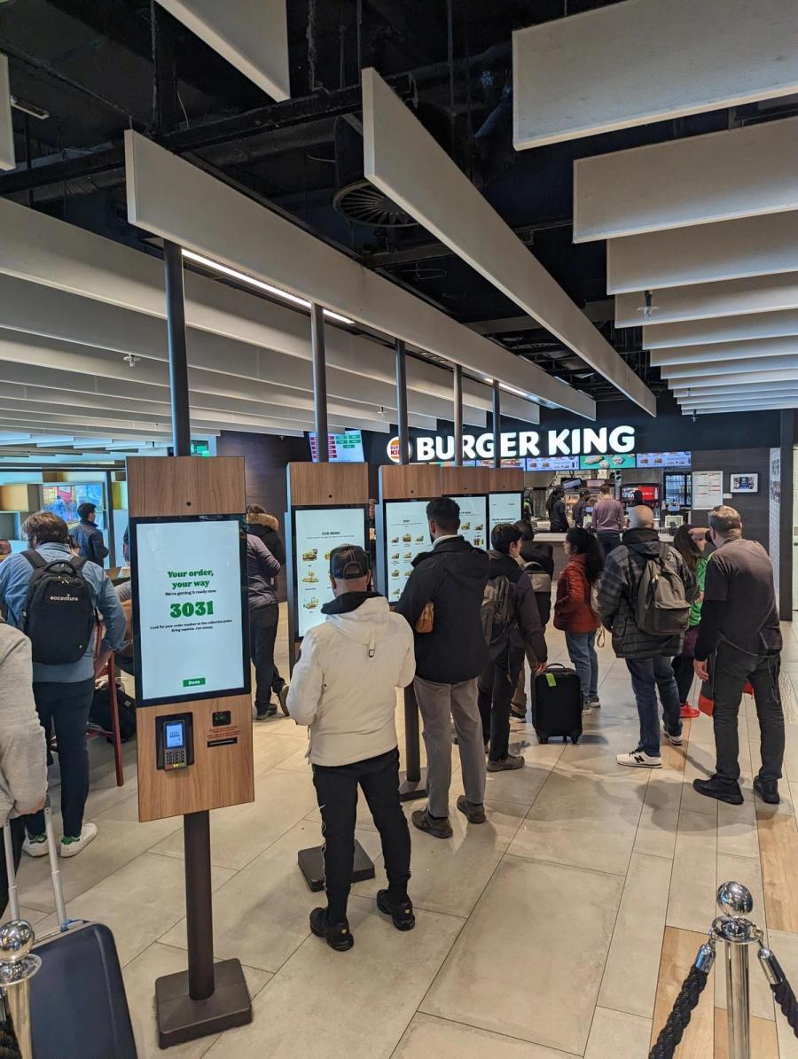 A row of Burger King self-ordering kiosks in a busy concourse, with guests queuing to order
