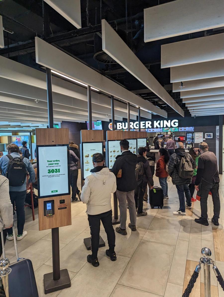 A row of Burger King self-ordering kiosks in a busy concourse, with guests queuing to order