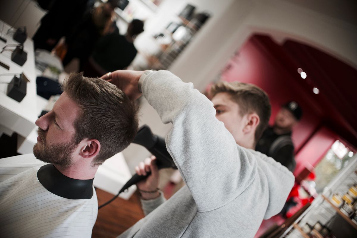 A barber styling a seated customer's hair with another barber behind, in front of a red wall
