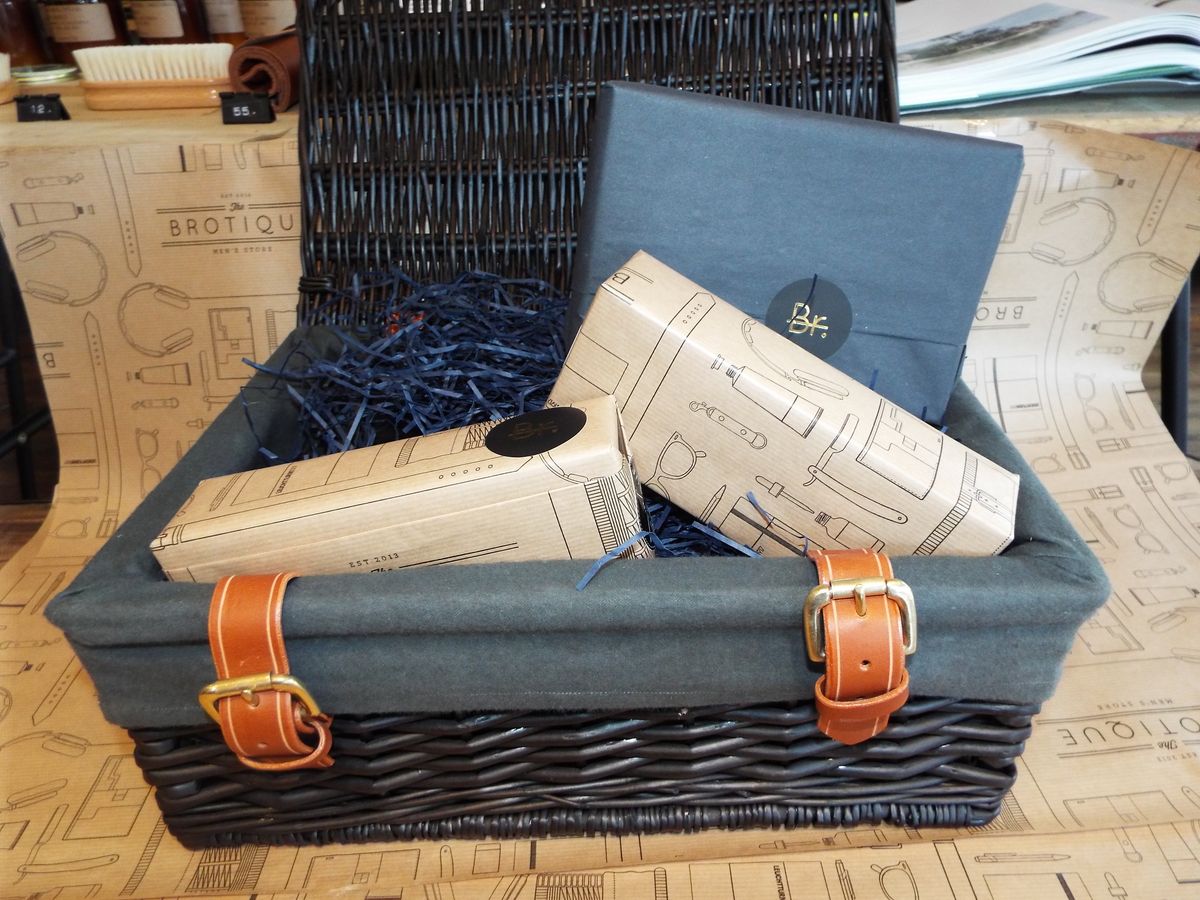 A wicker hamper lined with navy fabric and leather-buckled straps, holding gifts wrapped in Brotique paper and a navy box with a 'Br.' sticker