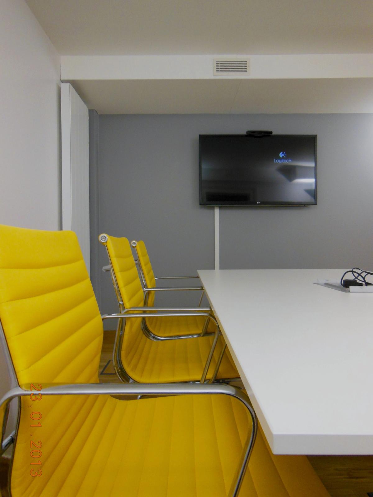 A white meeting room with yellow upholstered chairs around a white table and a wall-mounted screen