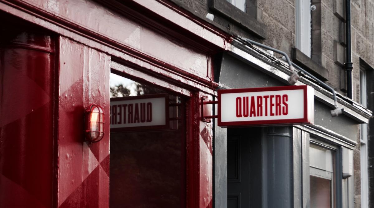 Close-up of a red-painted shopfront with a projecting illuminated sign reading 'QUARTERS' and a wall lamp