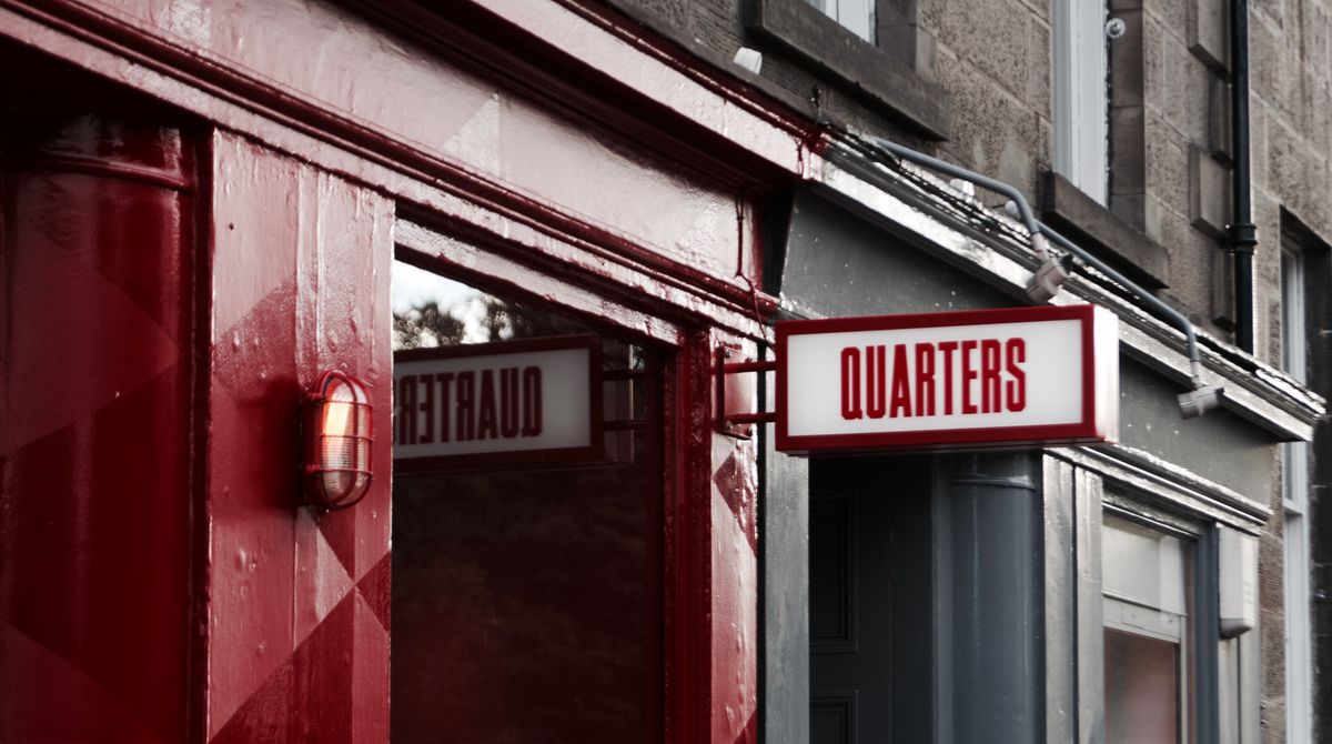 Close-up of a red-painted shopfront with a projecting illuminated sign reading 'QUARTERS' and a wall lamp