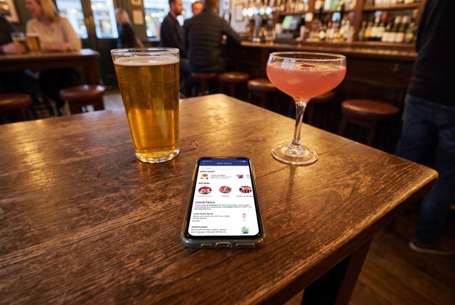 A phone lying on a wooden pub table between a pint of lager and a pink cocktail, showing a pub group's order-at-table menu with Order again, Our menu and Cocktails sections