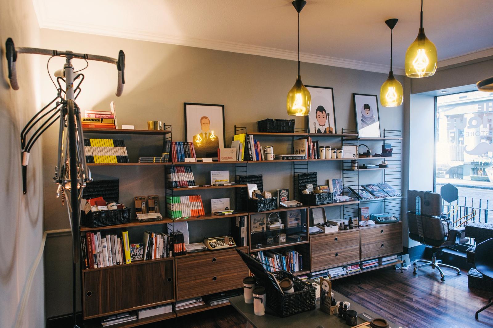 Interior of The Brotique: walnut and black wall shelving stocked with books, grooming products and accessories, framed illustrated portraits above, copper pendant lights and a road bike hung on the wall