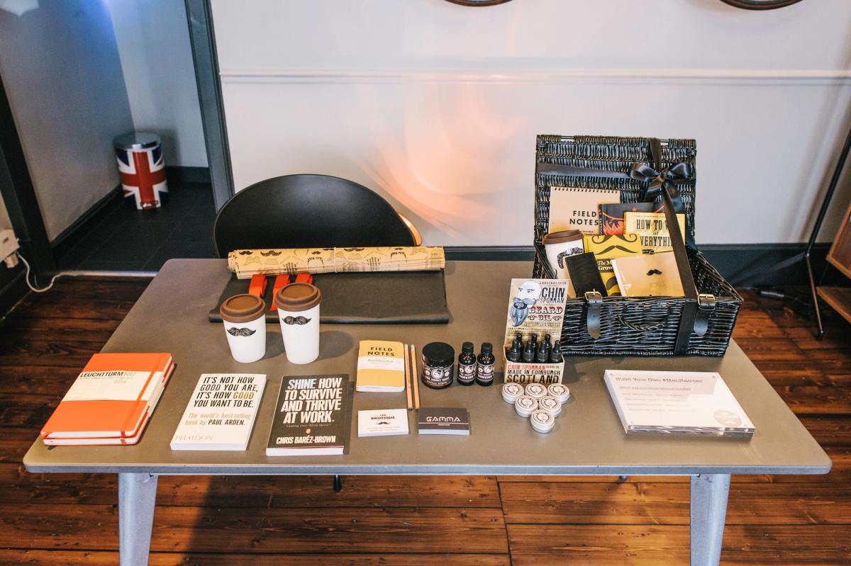 A grey display table on a dark wooden floor, laid out with notebooks, books, branded coffee cups, beard oil bottles and a basket of products