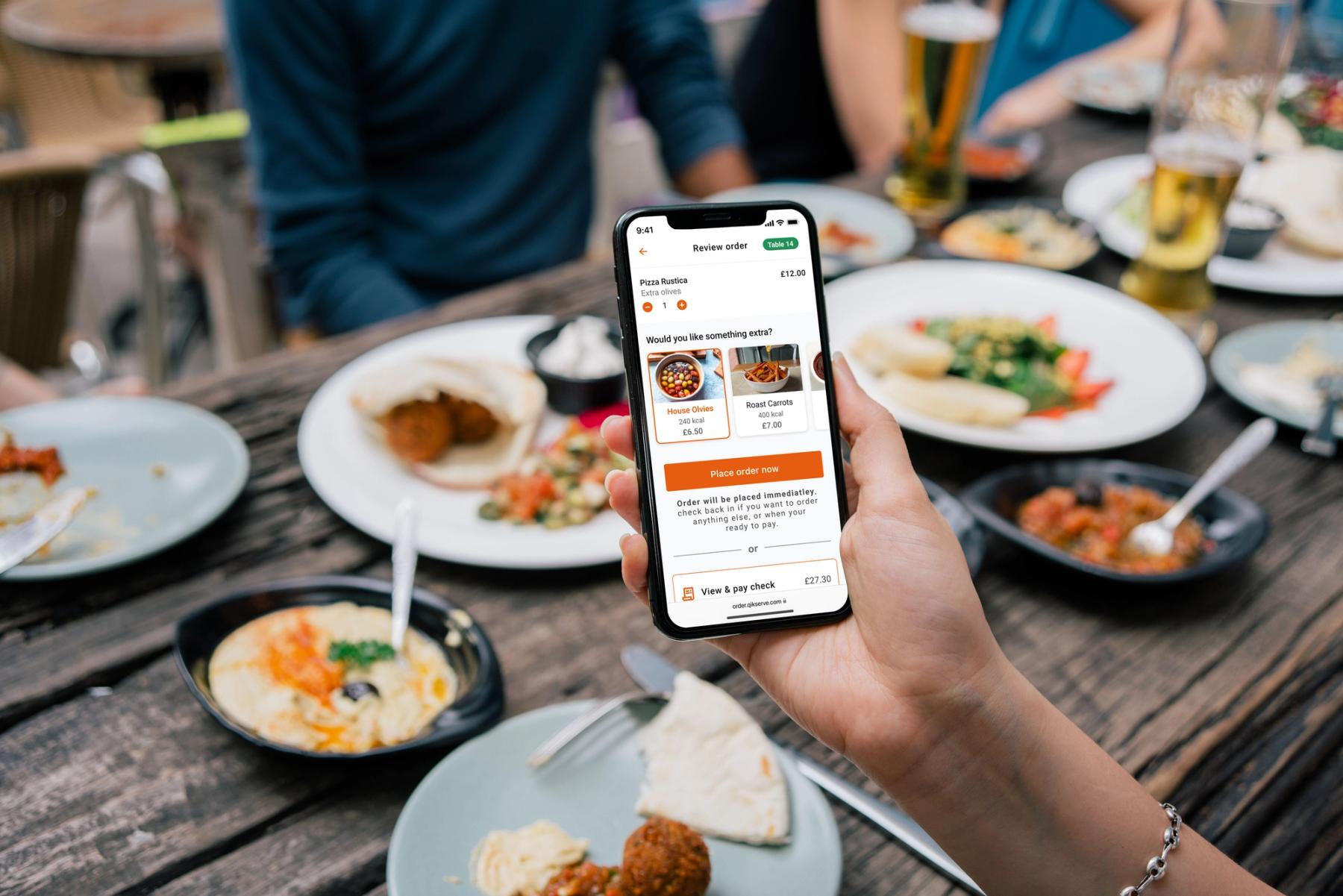 A hand holding a phone over a restaurant table of shared dishes and drinks, the phone showing an ordering and bill screen