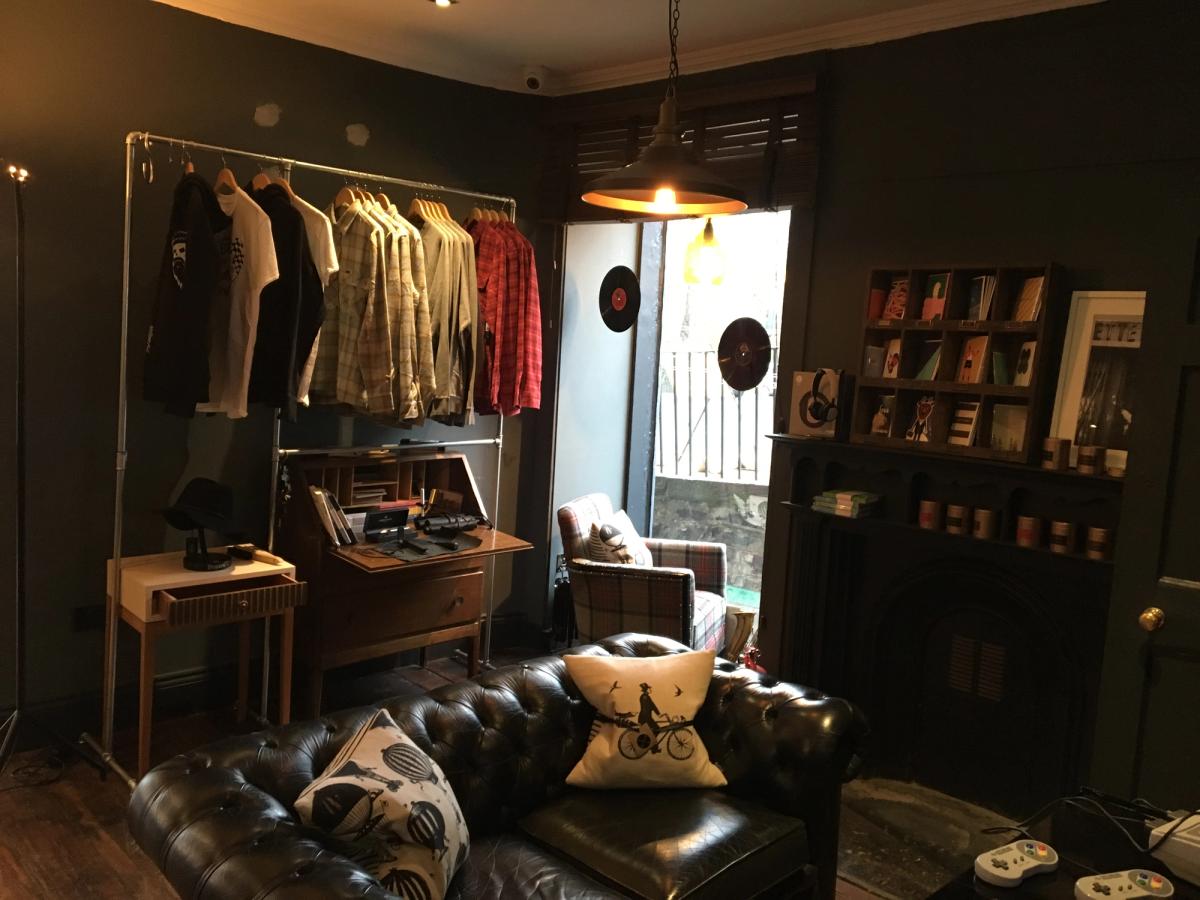 Corner of the Queen Street shop styled as a lounge: a black leather Chesterfield sofa, a clothing rail, a writing bureau and shelving under a pendant lamp