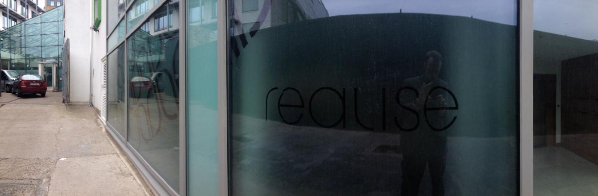 Panoramic street view of a ground-floor office frontage with dark glazing carrying a large frosted 'realise' logo
