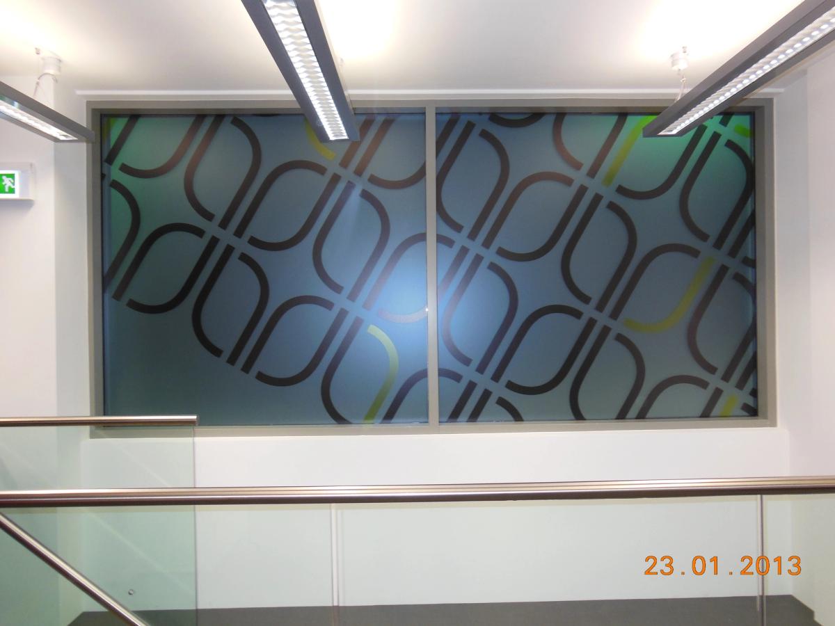 Office corridor glazing with a large frosted-film graphic of interlocking rounded shapes in grey and dark blue, under strip lights