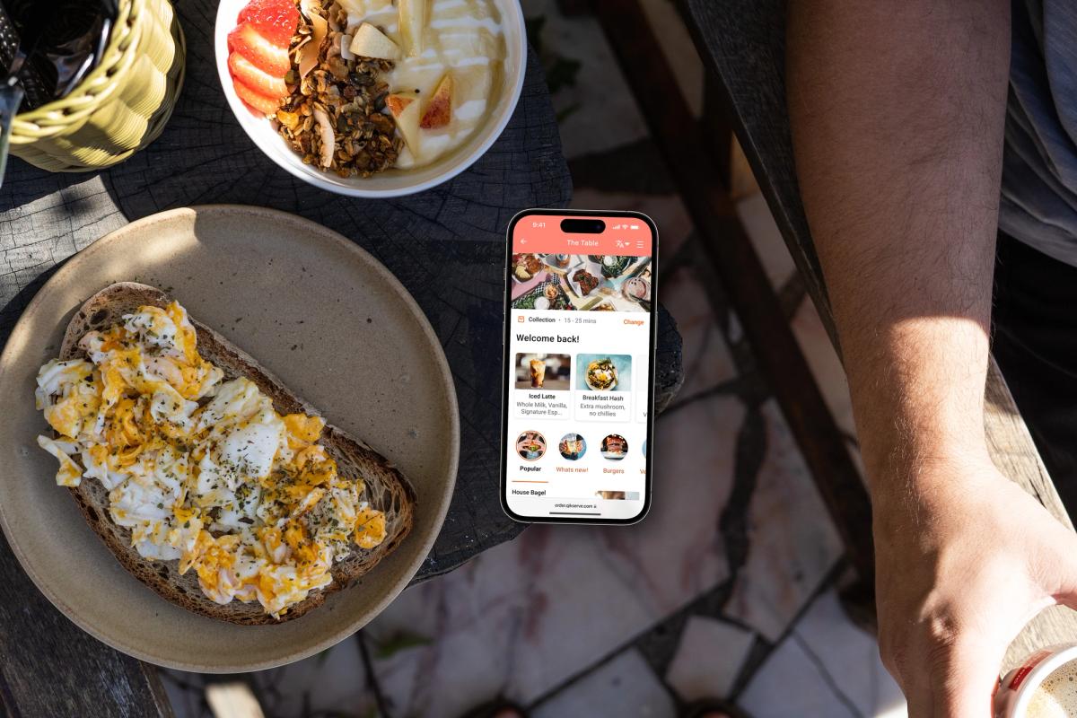 Overhead view of a breakfast table with eggs on toast and granola, and a hand beside a phone showing an ordering app home screen with an order-again row