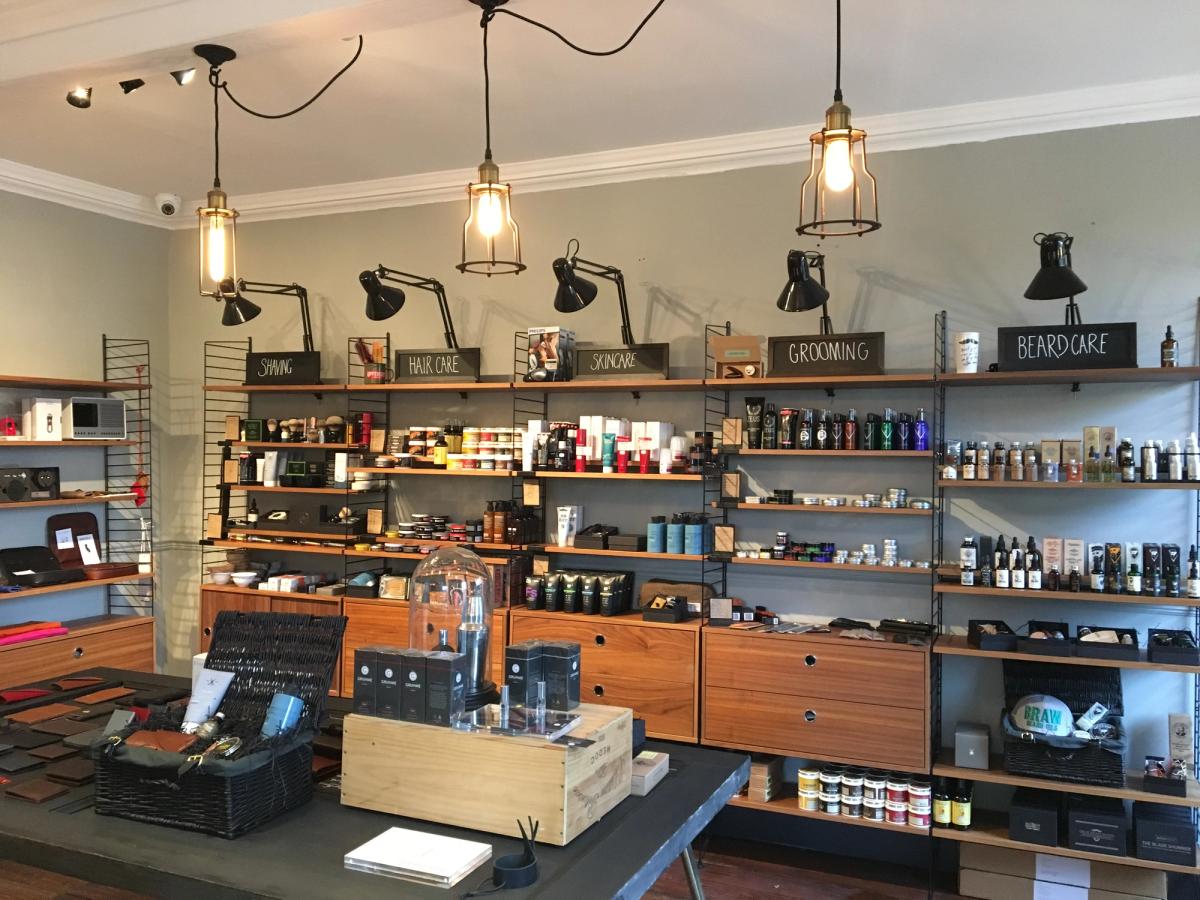 The Queen Street shop floor: a long run of wall shelving packed with grooming products under labelled sections, black task lamps and caged pendant bulbs, and a display counter