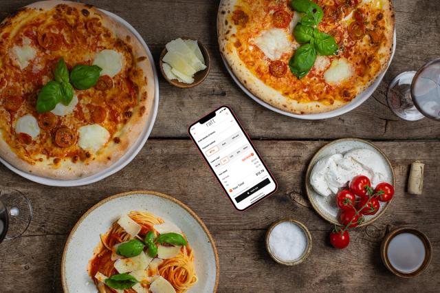 Overhead view of a rustic wooden table with pizzas, a bowl of pasta and small dishes, a phone lying among them showing an order and bill screen