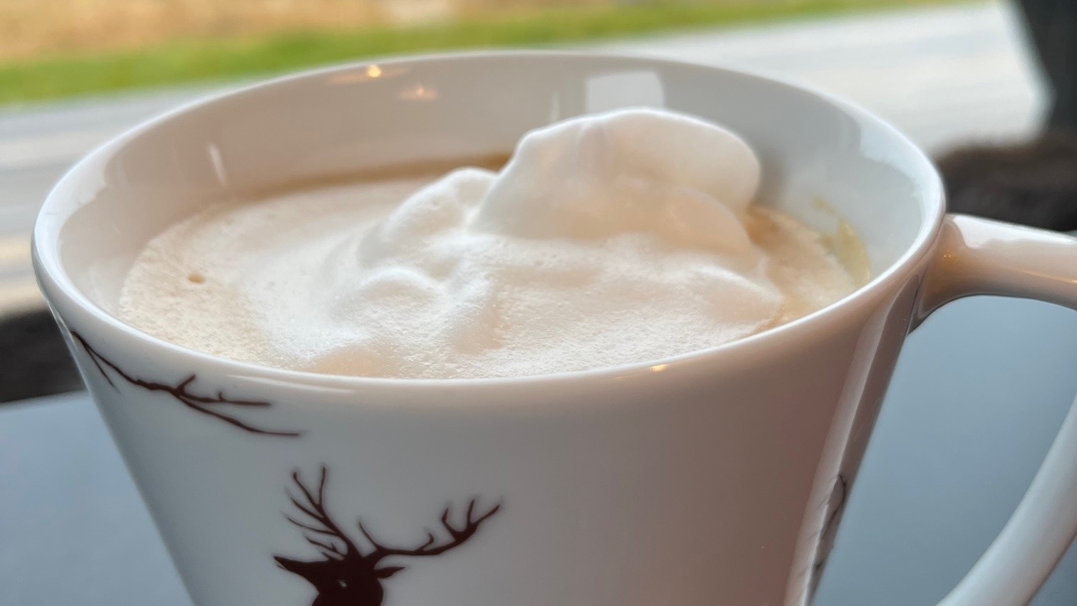 Kopp med bilde av en hjort står foran et vindu. Koppen er fylt med drikke med krem.