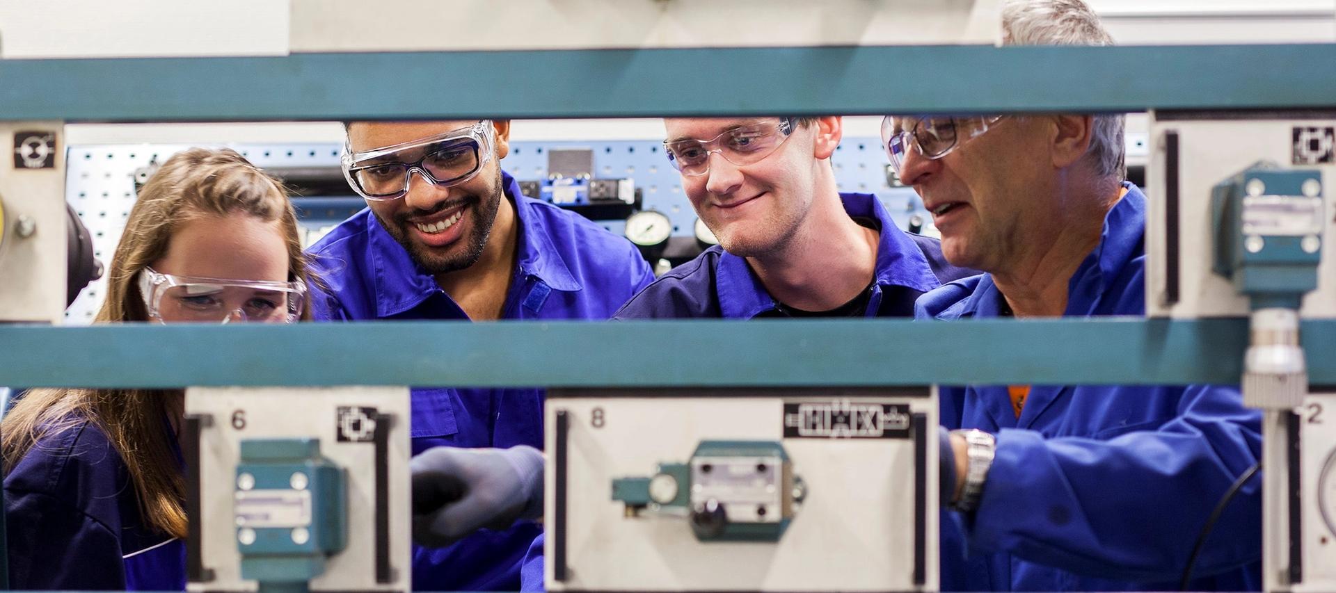 Nærbilde av 3 studenter og 1 lærer med vernebriller blå kjeledresser ser på en teknisk installasjon.