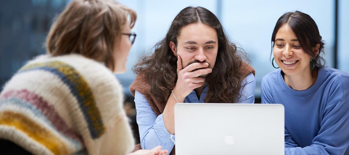 Tre studenter lener seg over en laptop. Den ene har ryggen til, én ser ettertenksom ut, den tredje smiler.