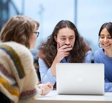 Tre studenter lener seg over en laptop. Den ene har ryggen til, én ser ettertenksom ut, den tredje smiler.