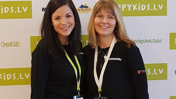 To kvinner smiler mot kamera mens de poserer foran en fotovegg på en konferanse.