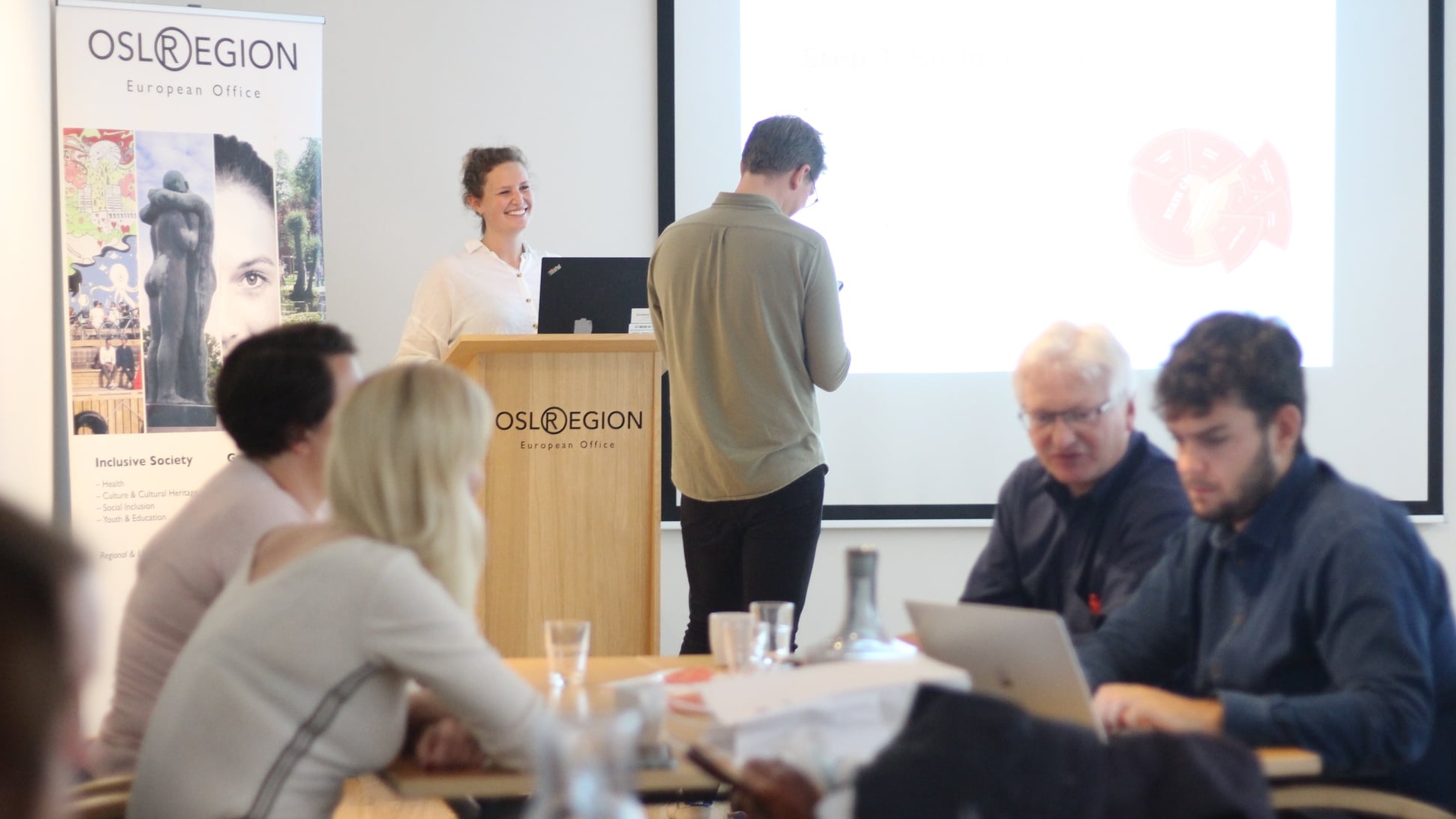 Kvinne står på talerstol i et møterom. Foran talestol står en mann og ser på en mobil, mens fire personer sitter rundt et bord.