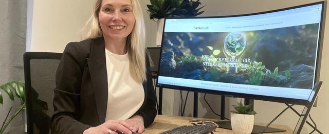 Alfhild Heyerdahl Krokan sitter ved et møtebord med PC og dokumenter foran seg. I bakgrunnen vises en skjerm med MerkeKraft-nettsiden.