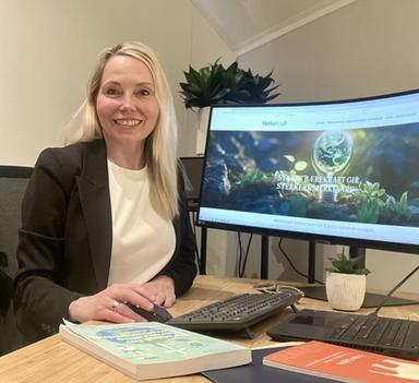 Alfhild Heyerdahl Krokan sitter ved et møtebord med PC og dokumenter foran seg. I bakgrunnen vises en skjerm med MerkeKraft-nettsiden.