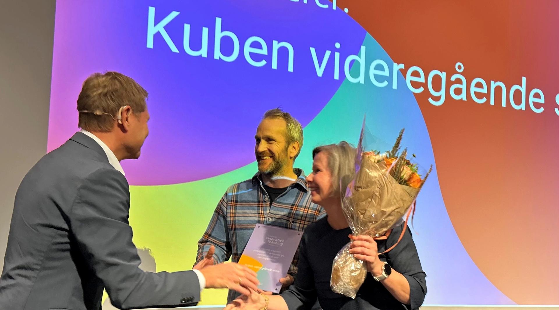 En mann deler ut blomster til to prisvinnere på en scene.
