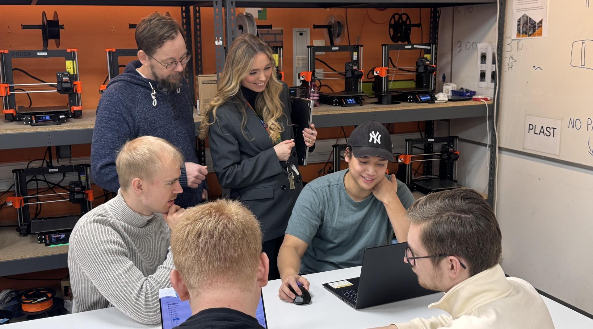 Fire studenter og to ansatte ved Høgskolen i Østfold samarbeider rundt en bærbar PC i et verksted med 3D-printere i bakgrunnen.