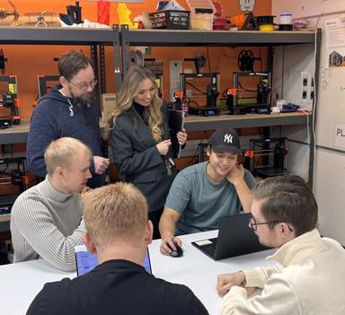 Fire studenter og to ansatte ved Høgskolen i Østfold samarbeider rundt en bærbar PC i et verksted med 3D-printere i bakgrunnen.