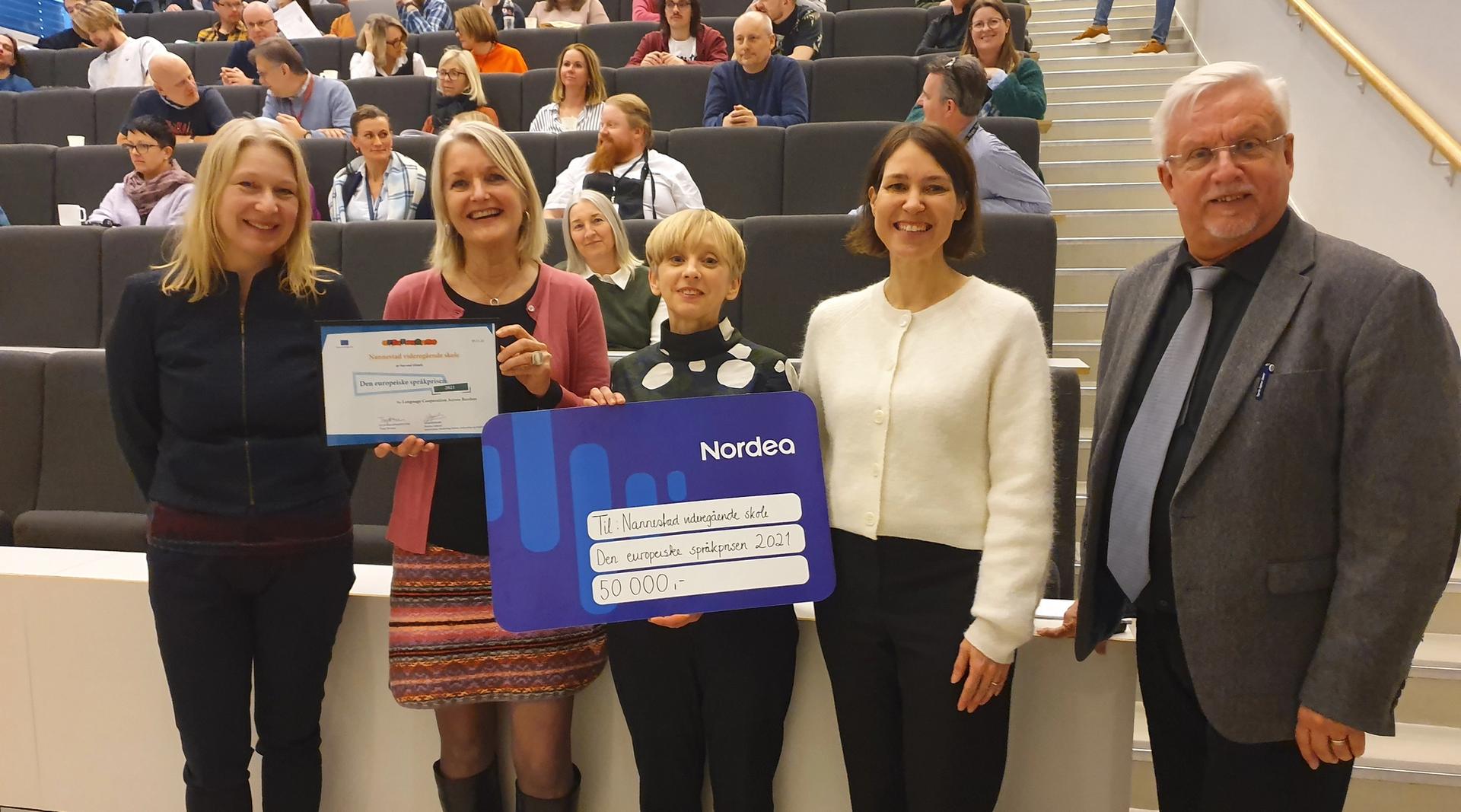 Tre kvinner, en mann og en gutt holder opp et diplom og en gavekort foran et fullsatt auditorium.