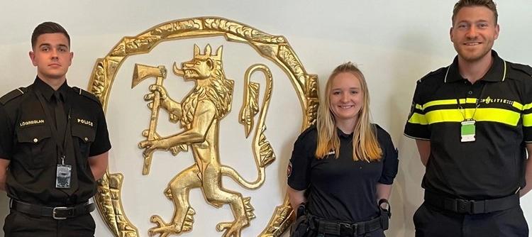 To menn og en kvinne står foran riksvåpenet. Smiler mot kamera.