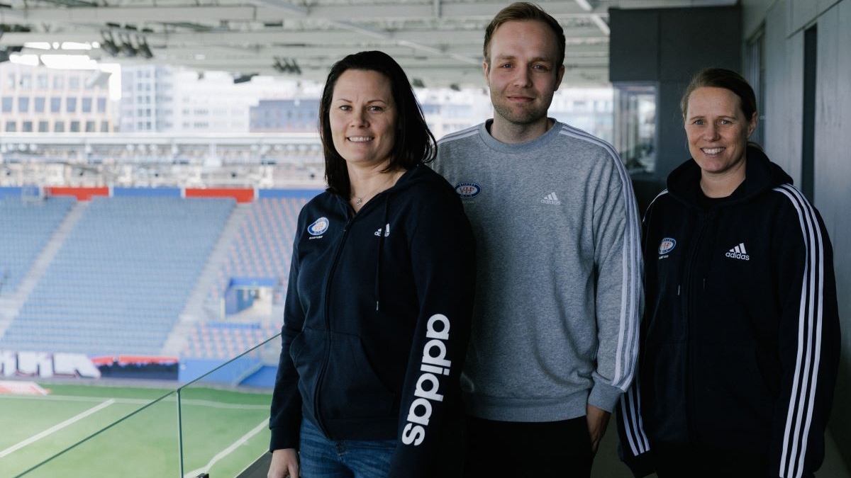 To kvinner og en mann smiler mot kamera. Står på en fotballtribune med fotballbanen i bakgrunnen.